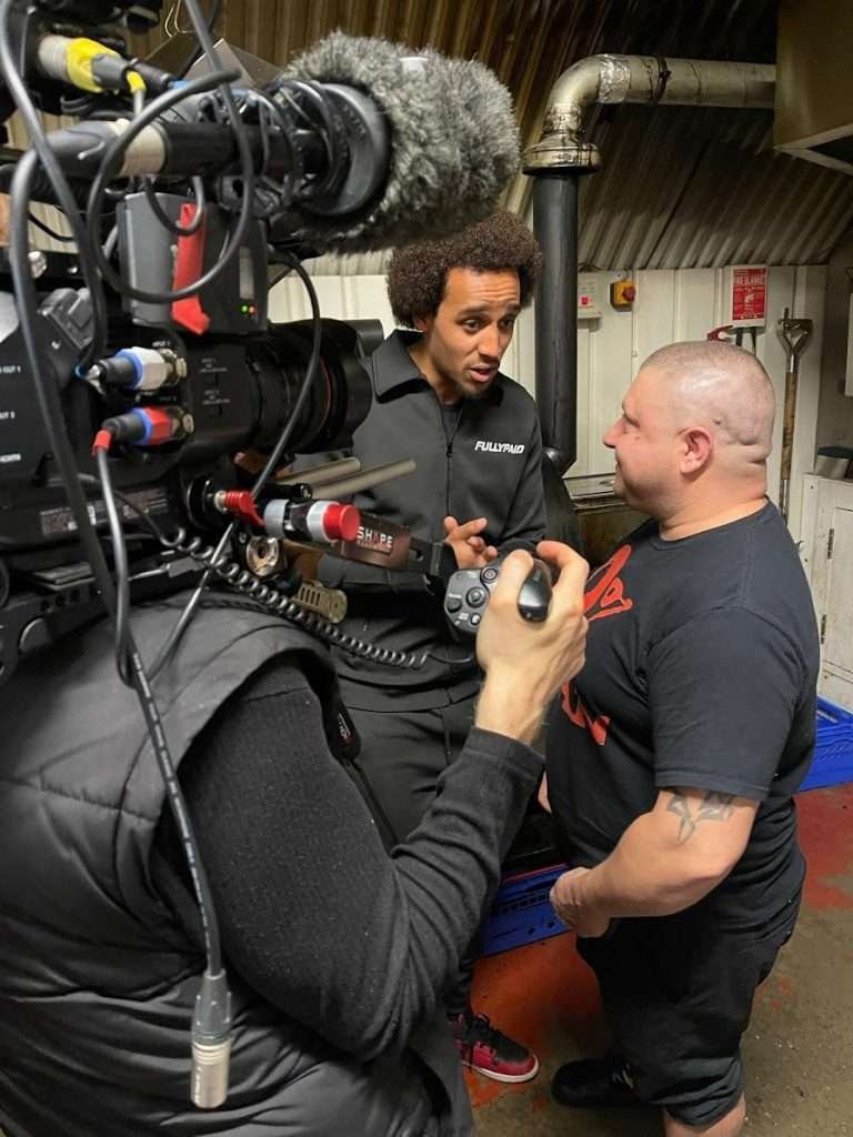 Behind the scenes photo of Omah talking with smoke house chef about cooking vegetarian food. Camera man is close to them with camera on his shoulder.