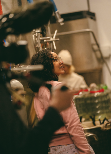 A young woman smiling and standing in a production setting, possibly a video shoot, wearing a pink hoodie. The scene includes filming equipment and a blurred person in the background. This image reflects creative content production for branding.