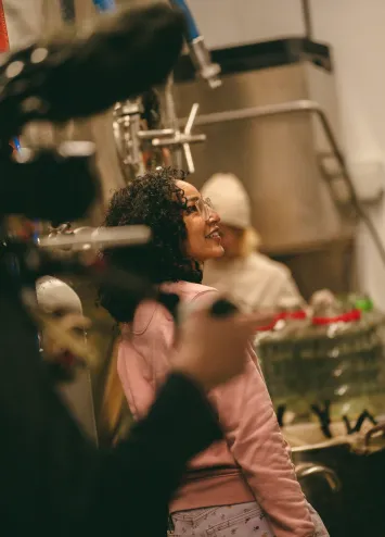 A young woman smiling and standing in a production setting, possibly a video shoot, wearing a pink hoodie. The scene includes filming equipment and a blurred person in the background. This image reflects creative content production for branding.