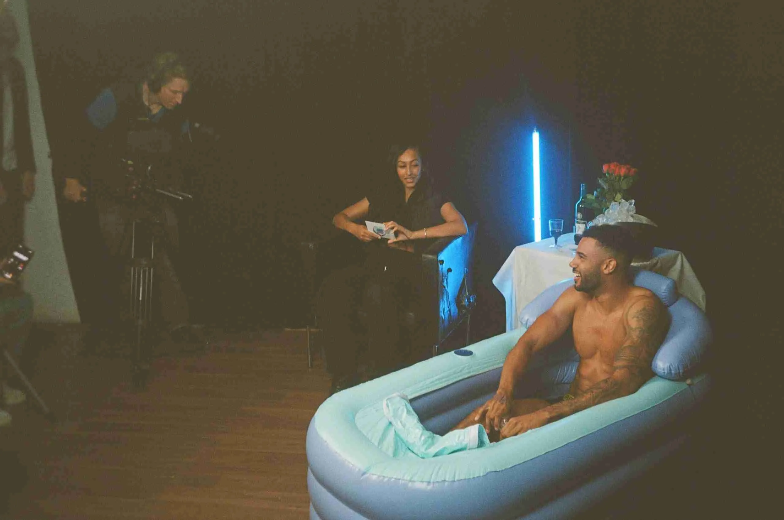 Video production set with a man in an inflatable pool being filmed. A woman is seated nearby with notes, and a cameraman adjusts the camera. Creative video shoot concept for brands.