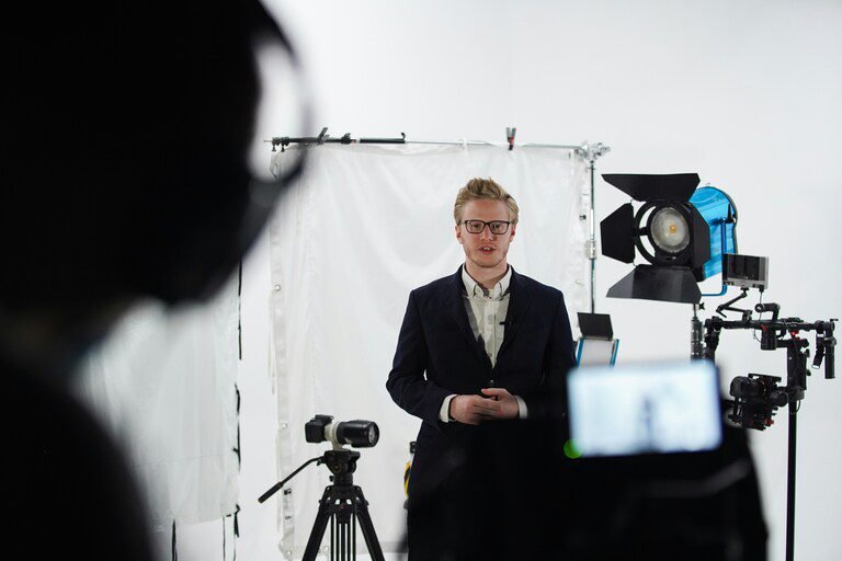A behind the scenes of a simple corporate video production. A contributor stood in front of camera with a white background waiting for lights and action.